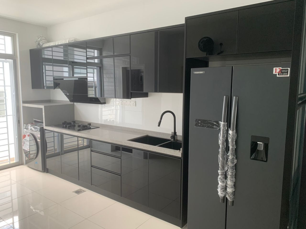 A long galley kitchen with grey gloss doors and black trim, a sink under the window, cooker hood, washing machine and a dark fridge.